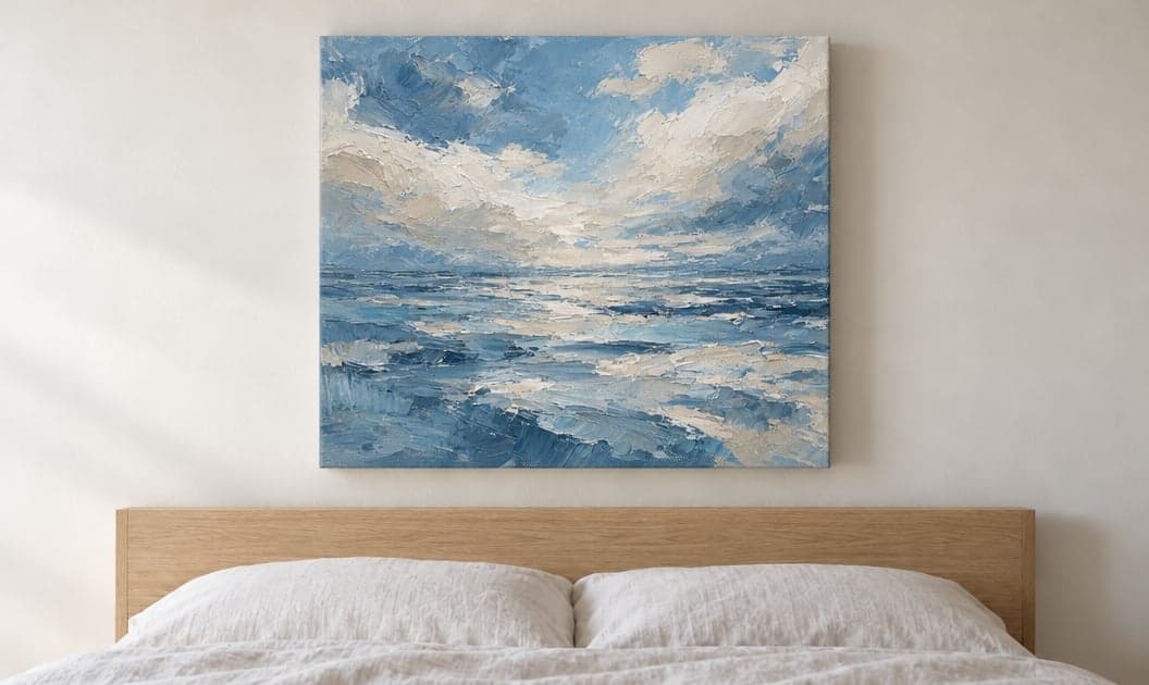 A canvas of a blue sea and clouds above a bed, hung without a frame